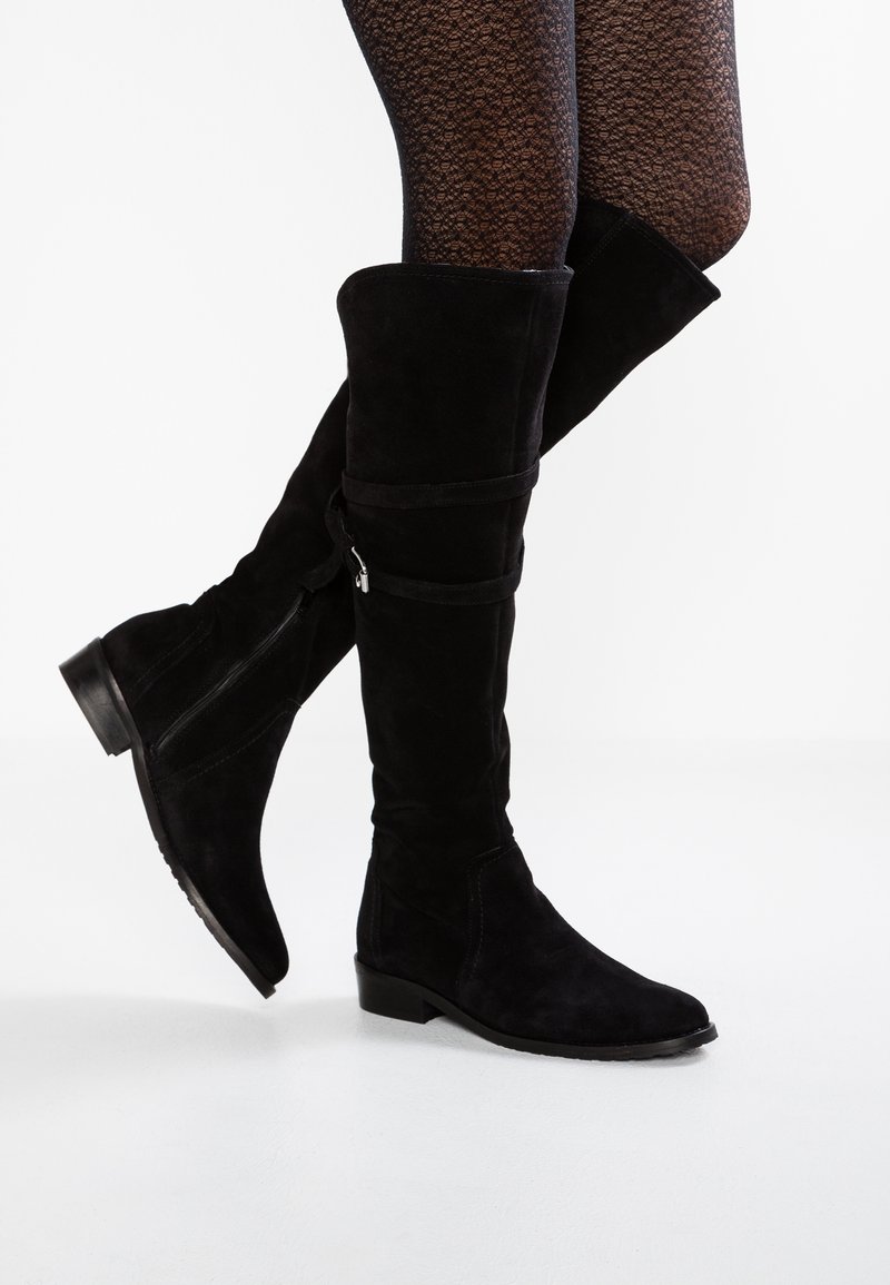 Botas de ante negras hasta la rodilla con tacón plano, que presentan una cremallera lateral y un detalle de correa en contraste alrededor de la pantorrilla. Acabado texturizado visible.