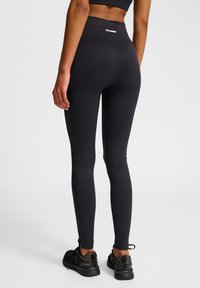 Czarne legginsy o prążkowanej fakturze i wysokim pasie; metka z logo z tyłu; zestawione z czarnymi butami sportowymi.