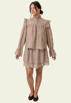 Frau in einem beigen Spitzenkleid mit langen Ärmeln und Rüschen an Schultern und Manschetten, steht und schaut seitlich, dunkle Haare hochgesteckt.