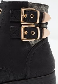 Gros plan d'une botte en cuir noir avec deux boucles en métal doré et un panneau texturé gris foncé près de la cheville.