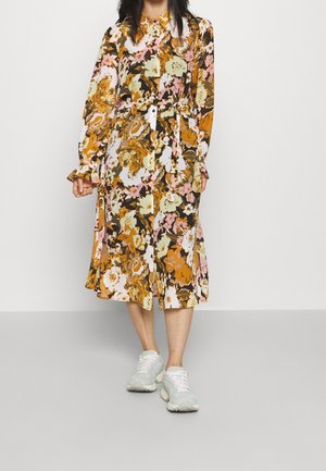 Femme portant une robe longue à manches longues à fleurs avec des boutons et une ceinture à la taille, associée à des baskets blanches, se tenant devant un fond uni.