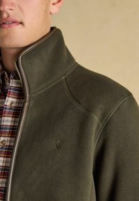 Fleeceová bunda v olivovej zelenej farbe s vysokým golierom a zipsom, na ktorom je jemné logo. Nosená cez károvanú košeľu. Mäkká textúra.