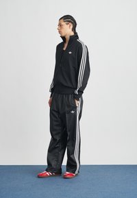 Čierna tepláková súprava z hladkého polyesteru s bielymi tromi pruhmi. Nohavice sú voľné; na nohách sú červené tenisky s bielymi pruhmi.