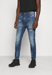 Jeans de mezclilla azul oscuro con un diseño de corte ajustado, que presentan desgastes, múltiples bolsillos y cierre de cremallera. Combinados con zapatillas deportivas negras.