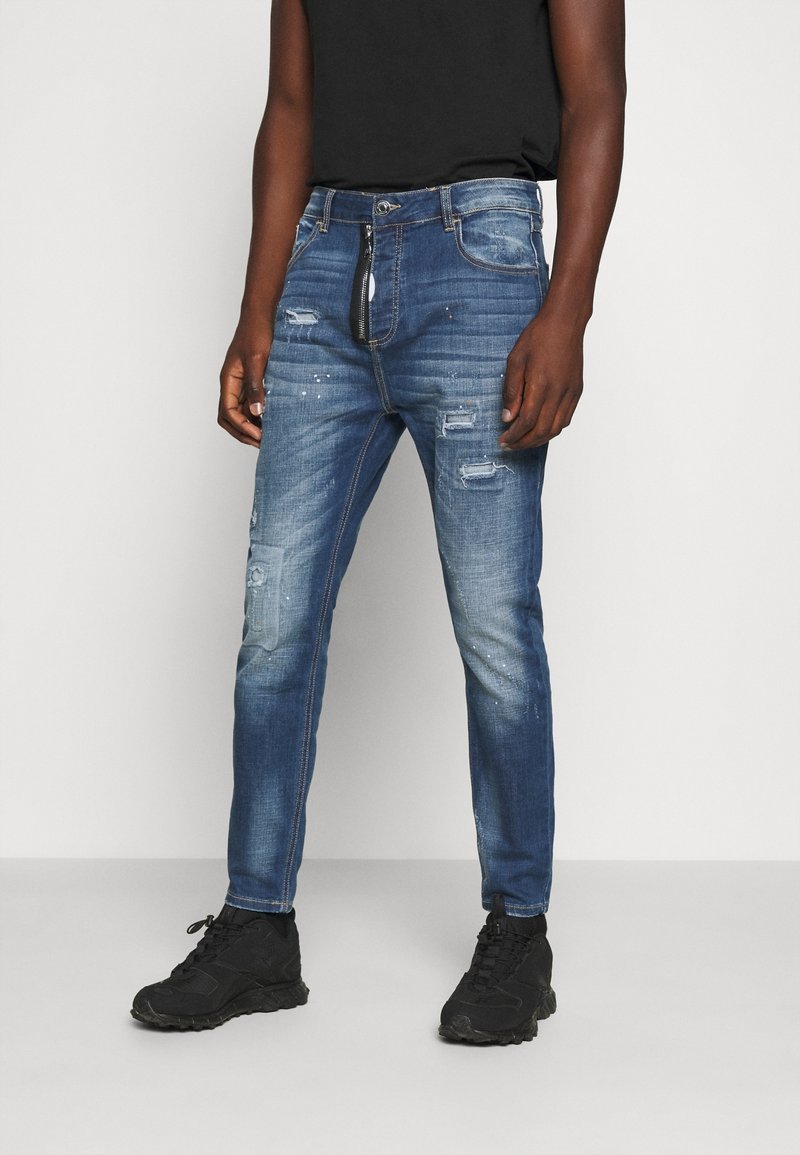 Jeans de mezclilla azul oscuro con un diseño de corte ajustado, que presentan desgastes, múltiples bolsillos y cierre de cremallera. Combinados con zapatillas deportivas negras.