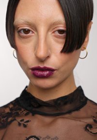 Primo piano di un volto con capelli corti e dritti di colore scuro. I tratti includono una pelle liscia, sopracciglia chiare e rossetto bordeaux intenso, abbinati a un top in pizzo nero.