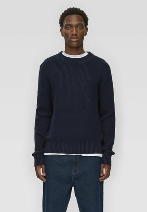 Navy ribbed knit sweater with a crew neckline, long sleeves, and textured fabric; worn over a white shirt, paired with dark denim pants.