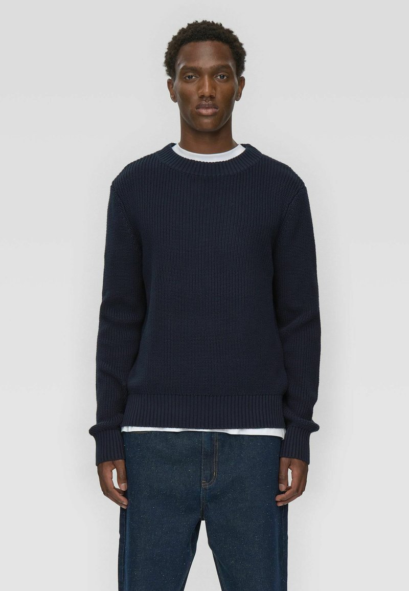 Pull en maille côtelée bleu marine avec un col rond, des manches longues et un tissu texturé ; porté par-dessus une chemise blanche, associé à un pantalon en denim foncé.