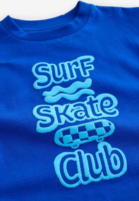 Sininen puuvillapaita, jossa on kohotettu vaaleansininen teksti "Surf", aaltoileva viiva, "Skate", ruudullinen skeittigrafiikka ja "Club" leikkisällä fontilla.