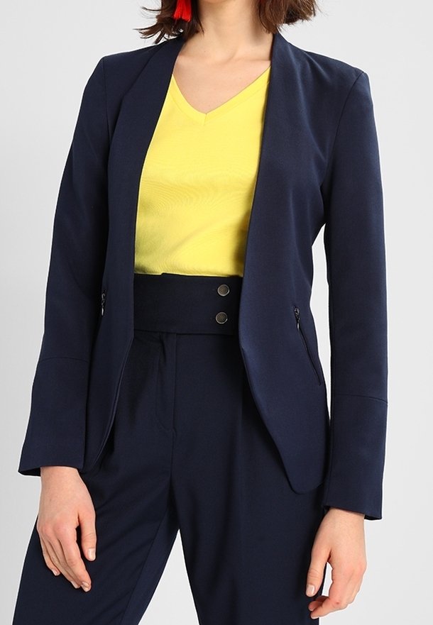 Femme portant un blazer ajusté bleu marine et un pantalon taille haute avec un chemisier jaune vif à col en V, et des boucles d'oreilles à pampilles rouges.
