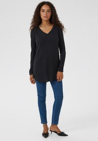 Black, long-sleeve tunic with a V-neck and henley-style placket, paired with blue slim jeans and black heeled mules. Smooth texture.