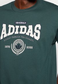 Camiseta verde de Adidas con "Originals Adidas World Famous for Quality 1972-2022" y un logotipo circular que celebra 50 años.