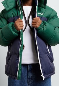 Zelená a tmavě modrá prošívaná bunda s předním zipem, vysokým límcem a pružnými manžetami. Obsahuje bílé zipové detaily a logo na prsou.