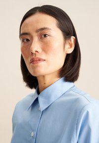 Camicia colletto blu chiaro realizzata in tessuto morbido, con chiusura a bottoni, design strutturato e una leggera lucentezza.