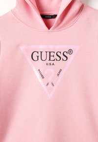 Różowa bluza z kapturem z trójkątnym logo, z napisem "GUESS U.S.A." w czerni, otoczonym błyszczącym jasnoróżowym obramowaniem. Materiał bawełniany, styl casual.