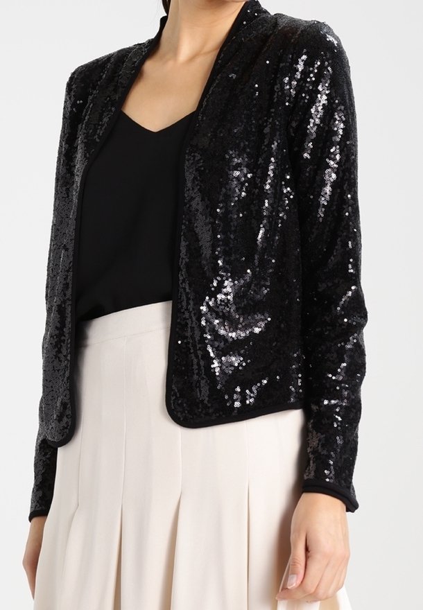 Femme portant une veste noire à paillettes ouverte devant sur un haut noir et une jupe plissée crème taille haute.