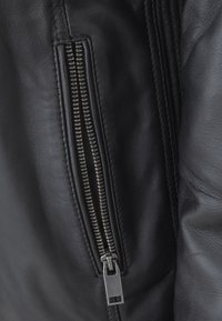 Giacca di pelle nera con una cerniera metallica, caratterizzata da una superficie liscia e dettagli cuciti lungo la cucitura. I tirazip sono in argento.
