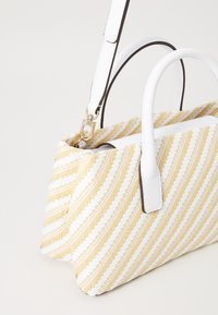 Le sac en toile présente un motif rayé diagonal jaune et blanc. Sa forme structurée est dotée de poignées synthétiques blanches et d'une sangle amovible.