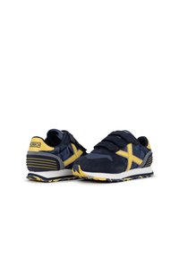 Zapatillas azul marino con acentos amarillos, parte superior de ante y tela, correas de gancho y lazo, suela texturizada y detalle de patrón geométrico.