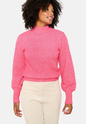 Roze gebreide trui met een hoge hals, ruitpatroon en puffmouwen. Geribbelde zoom en manchetten, gecombineerd met lichtgekleurde geribbelde broeken.