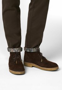 Bruine suède enkellaarsjes met een ronde neus, voorzien van gouden hardware en een textiel manchet met een patroon. G gedragen met een bruine broek.