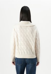 Personne avec des cheveux foncés et droits portant un pull en tricot torsadé crème et un jean bleu, de dos contre un fond blanc.