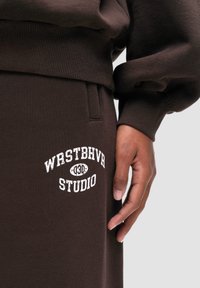 Brauner Pullover mit Rippbündchen und -saum, kombiniert mit passender brauner Jogginghose mit weißem "WRSTBHVR STUDIO" Textlogo.