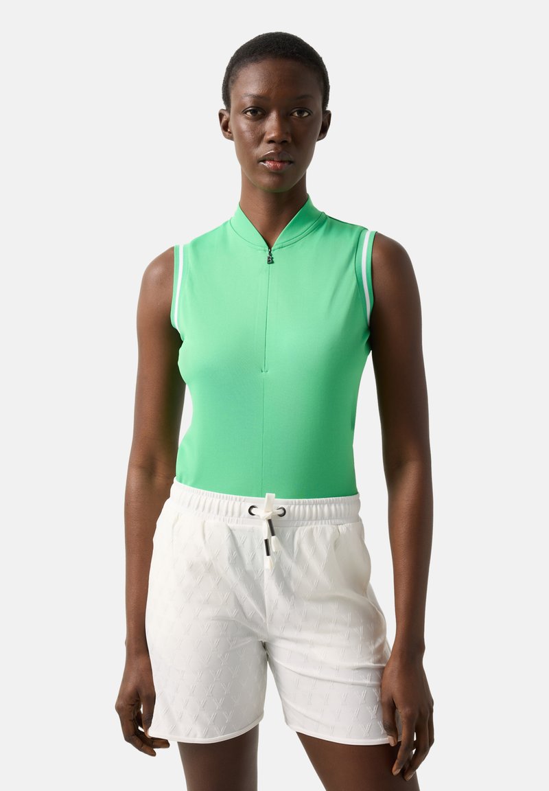 Vrouw met een mouwloos groen topje met rits en witte shorts met trekkoord, staand tegen een eenvoudige lichte achtergrond.