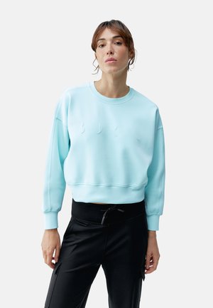 Svetlo modra skrajšana bluza z okroglim izrezom, reberčastimi manšetami in vrezanim logotipom na sprednji strani. Usklajena s črnimi hlačami.