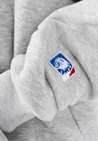 Tela gris claro con un puño acanalado y un pequeño parche bordado de una pantera rugiente en azul, blanco y rojo en la manga.