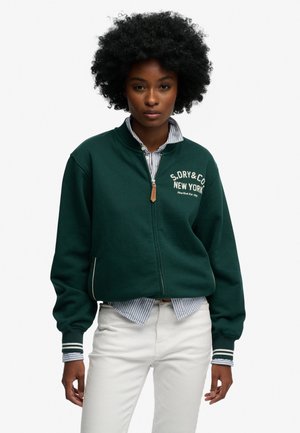 Zielona bluza zapinana na zamek z prążkowanymi mankietami i kołnierzem, z haftowanym białym logo. W komplecie z niebieską i białą w paski koszulą pod spodem.