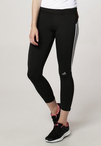 Černé sportovní legíny s bílými bočními pruhy, přiléhavý střih, pružný materiál a logo Adidas na spodní části nohy.