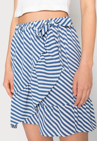 Saia mini azul e branca com riscas, apresentando um design envolvente, com um laço na cintura, tecido texturizado e uma bainha em franzido.