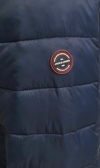 Tamsiai mėlyna pūkinė striukė su apvaliu juodu ir raudonu logotipo pleistru, ant kurio užrašyta "JACK & JONES", ir tekstūruotais horizontaliais siūlais.