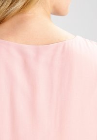 Blouse rose clair avec une texture douce et lisse, dotée d'un large décolleté, présentant une coupe décontractée et des détails de design minimaux.