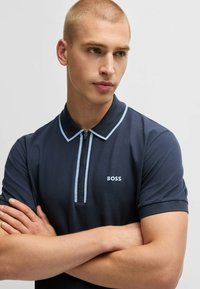 Polo in navy con accenti azzurri, colletto zip e un piccolo logo bianco. Il tessuto sembra liscio e aderente.