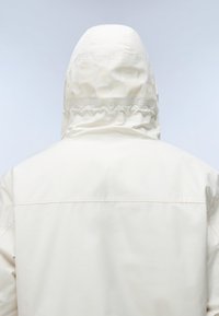Rückenansicht einer Person, die eine weiße Kapuzenjacke mit elastischem Detail an der Rückseite der Kapuze vor einem hellgrauen Hintergrund trägt.