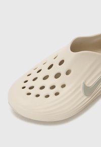 Beige sleehattar med en slät finish, med flera cirkulära utskärningar och en silverfärgad Nike-logotyp som accent på sidan.