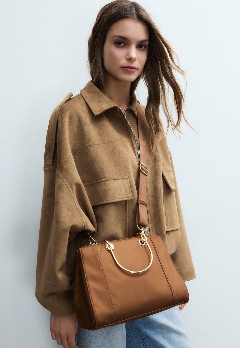 Jeune femme en veste en daim marron portant un sac à main en cuir beige avec des poignées dorées, vêtue d'un jean bleu, sur un fond clair uni.