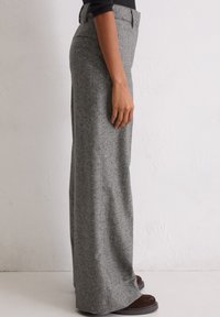 Persoon in een hoge taille, met wijde grijze broek en donkerbruine schoenen, staande in profiel tegen een effen witte muur.