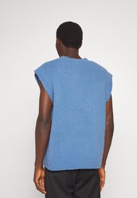 Personne à la peau foncée portant un gilet bleu sans manches en maille ample et un pantalon noir, vue de dos sur un fond clair uni.