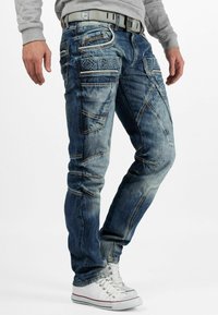 Jeans en denim bleu à la coupe ajustée, avec des coutures apparentes, de multiples poches et une texture légèrement usée.
