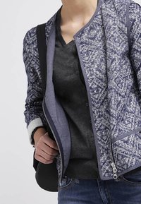 En person som bär en blå mönstrad zip-up jacka över en mörkgrå skjorta med knappar, blå jeans och bär en svart väska över axeln.