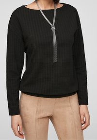Zwarte gestructureerde top met lange mouwen en een boot-halslijn, gecombineerd met tan suede-achtige broek. Een zilveren knoopketting voegt detail toe.