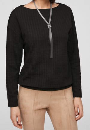 Haut noir texturé à manches longues avec un col bateau, associé à un pantalon beige en suède. Un collier en argent noué ajoute une touche de détail.
