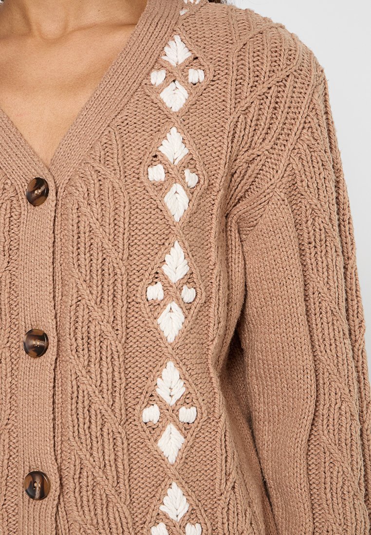 Beige Strickjacke mit weißen Rautenmuster, ausgestattet mit strukturierten Zopfnähten und braunen Knöpfen an der Vorderseite.