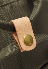 Grønn stoffreim med en lys brun lærflik sikret med en messingtrykkknapp. Teksturen inkluderer et vevd mønster og glatt lær.
