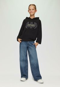 Zwarte hoodie met een vleermuislogo in een groen en wit gaaspatroon, gecombineerd met losse blauwe denim jeans en witte sneakers.