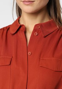Mujer vestida con una camisa de botón en color óxido, con cuello y bolsillos en el pecho, que muestra desde la barbilla hasta la mitad del pecho.