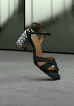 Black sandal with woven block heel, ankle strap with buckle, and crossed front straps, positioned on a gray surface with a reflective background.